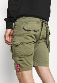 Pantaloni scurți cargo de culoare verde măslin, cu multiple buzunare, petice din țesătură și o curea detașabilă. Prezintă un eticheta roșie și un material texturat.