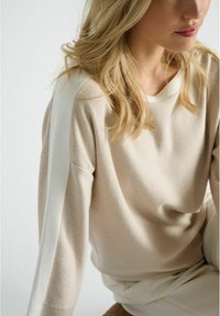 Pull en tricot beige avec une bande blanche le long des manches. Il présente une coupe décontractée et un col rond, la texture semble douce et lisse.