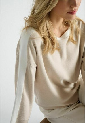 Beige gebreide trui met een witte streep langs de mouwen. Heeft een ontspannen pasvorm en een ronde halslijn, de textuur lijkt zacht en glad.