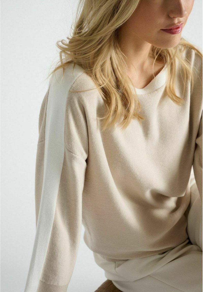 Pull en tricot beige avec une bande blanche le long des manches. Il présente une coupe décontractée et un col rond, la texture semble douce et lisse.