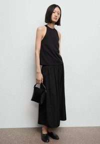 Top halter nero abbinato a una lunga gonna nera fluida. La modella tiene in mano una piccola borsa nera e indossa scarpe nere flat. Tessuto di texture liscia.