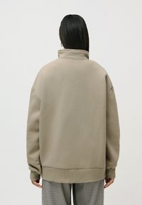 Person med langt svart hår iført beige høyhalset genser og svart-hvite rutete bukser, sett fra bakgrunnen mot en ensfarget bakgrunn.