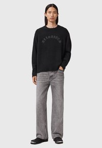 Man with long hair wearing black AllSaints sweater, gray wide-leg jeans, and black shoes, standing with one hand in pocket.