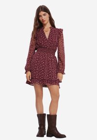 Robe florale bordeaux avec de longues manches transparentes, taille smockée, ourlet froncé et bottines marron. Présente un décolleté en V profond.