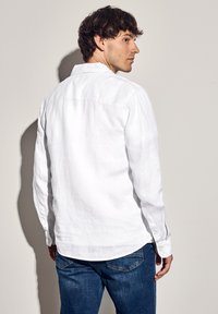Camisa blanca de lino de manga larga con cuello abotonado, dobladillo recto y puños con botones, combinada con vaqueros azules.