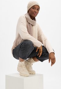 Gorro de punto beige y bufanda suave en tonos neutros combinados con botas claras de cordones beige. La modelo lleva una camiseta de manga larga clara y unos jeans cortos oscuros.