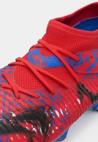 Chaussure de football rouge avec une texture en maille, dotée d'accents bleus et de motifs graphiques. Comprend des lacets rouges et un design de bout arrondi.