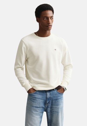 Bărbat purtând un pulover alb cu mâneci lungi și blugi albaștri, cu mâinile în buzunare, uitându-se într-o parte pe un fundal alb simplu.