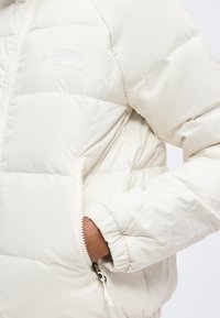 Χέρι στην τσέπη λευκού αφρωδούς μπουφάν North Face με ορατό φερμουάρ και ελαστική μανσέτα στο μανίκι.