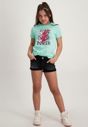 Mintgroene t-shirt met "GIRL POWER" en een rode tijgerafbeelding, gecombineerd met zwarte denim shorts en witte sneakers met oranje accenten.