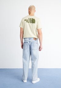 Camiseta de algodón beige con estampado gráfico verde en la parte posterior, combinada con jeans de mezclilla rectos azul claro y zapatillas blancas.