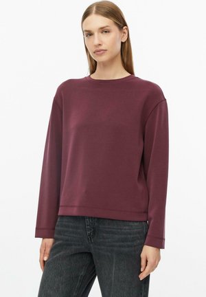 Femme aux cheveux raides châtain clair portant un haut bordeaux uni à manches longues et un jean gris foncé sur fond blanc.