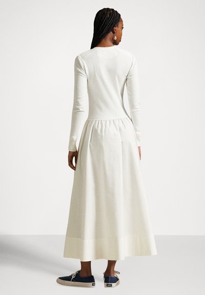 Μακρύ λευκό φόρεμα με μακριά μανίκια, εφαρμοστό κορσέ και φούστα με πιέτες. Συνδυάζεται με ναυτικό μπλε παπούτσια, το ύφασμα φαίνεται λείο και ελαφρύ.