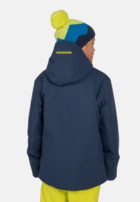 Giacca impermeabile blu navy con cappuccio, con un logo sulla schiena. Indossata con pantaloni gialli brillanti e un berretto lavorato a maglia multicolore.