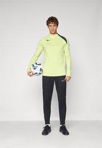 Odzież sportowa Nike ma jasną żółtą bluzę z długim rękawem, z czarnymi akcentami oraz czarne spodnie. Postać trzyma wzorzystą piłkę nożną.