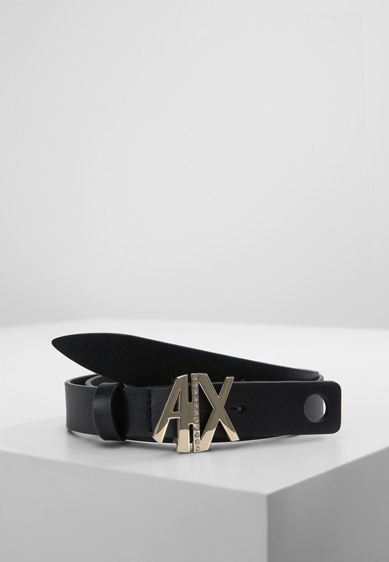 Ceinture en cuir noir avec boucle de logo métallique dorée "A|X" ornée de petits strass, enroulée sur une surface blanche.