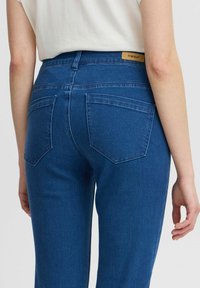 Tumedat sinised denimpüksid, millel on kõrge vöökoht, kaks tagumist taskut diagonaalse kujundusega ja nahkne brändilogo taga.