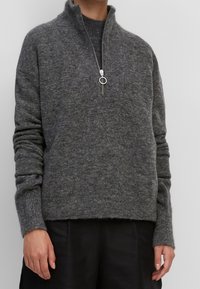 Persona che indossa un maglione di lana testurizzato grigio scuro con zip a metà e tiraglio ad anello, abbinato a pantaloni neri, in piedi contro uno sfondo neutro.