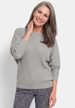 Femme d’âge moyen souriante aux longs cheveux gris portant un pull gris côtelé et un pantalon gris foncé à motifs, debout devant un fond clair.