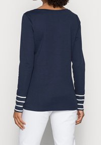 Marinblå långärmad T-shirt med randiga manschetter, rund halsringning och avslappnad passform. Tillverkad av mjuk bomull. Vita byxor synliga.