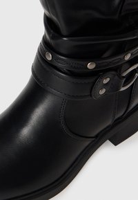 Bottine en cuir noir avec brides décoratives cousues et clous argentés, dotée d’un talon bas et épais et d’un bout rond.