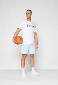 Hvit t-skjorte med svart logo, lyseblå mesh basketballshorts og hvite joggesko. Shortsene har et geometrisk mønster og detaljer med kant.