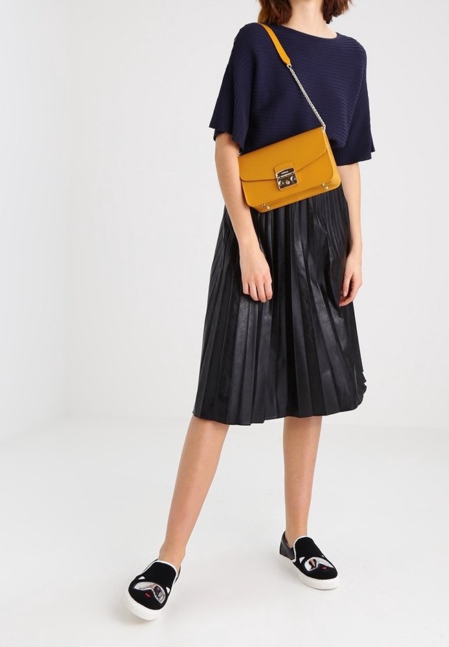 Woman wearing navy blue short-sleeve top, black pleated midi skirt, black slip-on shoes, and mustard yellow crossbody bag with gold clasp.