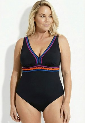 Frau trägt einen schwarzen Einteiler mit blauem, lila und orangefarbenem Zickzackbesatz am Ausschnitt und unter der Brust, stehend vor weißem Hintergrund.