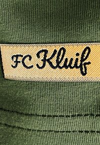 Groene gebreide muts met een gestructureerd oppervlak. Heeft een oranje label met zwarte tekst "FC Kluif." Eenvoudige en functionele ontwerpelementen.