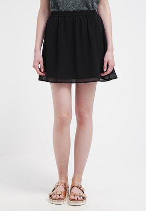 Vrouw die een zwarte rok met een elastische tailleband draagt, een grijze top en sandalen met een rosegouden gesp, staand tegen een effen witte achtergrond.