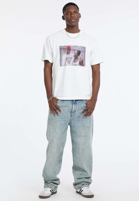 Biała bawełniana koszulka z graficznym nadrukiem przedstawiającym dwie osoby. Połączona z jasnoniebieskimi, luźnymi dżinsami i białymi trampkami z czarnymi akcentami.