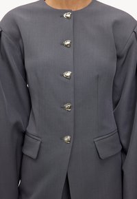 Blazer gris à encolure ronde, manches bouffantes, cinq boutons métalliques et deux poches. Tissu lisse avec une coupe ajustée.