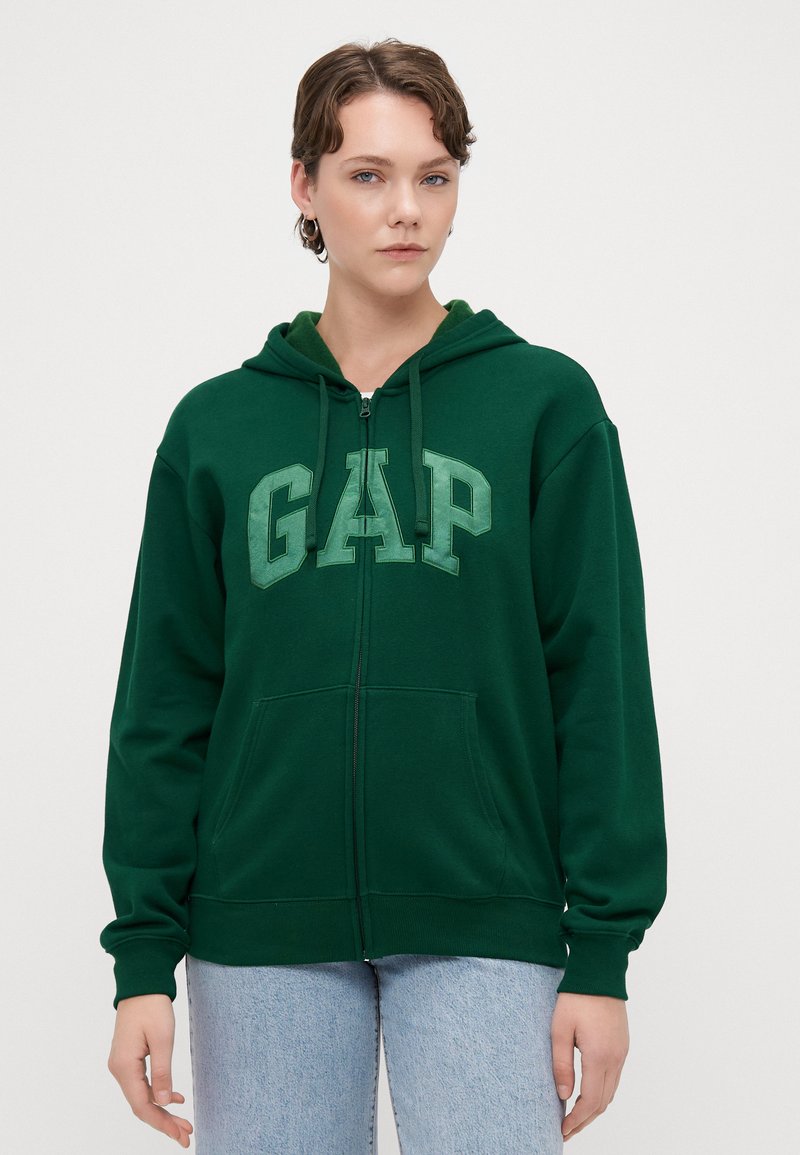 Sudadera verde con cremallera hecha de tela suave, con un gran logo "GAP" en un tono más claro. Incluye una capucha y bolsillos frontales.