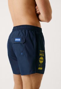Bañador de color azul marino hecho de tela ligera con cintura elástica, bolsillo frontal y logotipos amarillos impresos en el lateral y en la parte posterior.