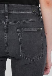 Gros plan sur une personne portant un jean gris foncé, montrant les détails de la poche arrière et une partie du bras droit contre un fond uni.