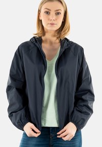 Donker marineblauwe, capuchon regenjas met elastische cuffs en een ritssluiting aan de voorkant. Lichtgewicht, glanzend materiaal. G gedragen over een lichtgroene top en blauwe jeans.