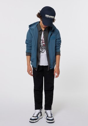 Blauwe hoodie-jack, grijze overhemd met knopen, witte graphic t-shirt, zwarte broek en zwart-witte sneakers met blauwe accenten.