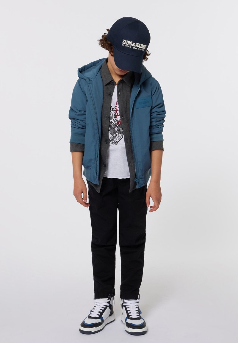 Veste à capuche bleue, chemise gris boutonnée, t-shirt graphique blanc, pantalon noir et baskets noir et blanc avec des accents bleus.