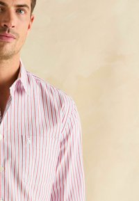 Biela košeľa na gombíky s červenými a modrými vertikálnymi pruhmi. Obsahuje predné vrecko a golier. Vyrobená z ľahkej tkaniny s hladkou textúrou.