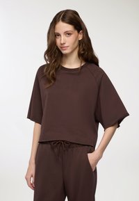Sweatshirt corto marrone con maniche ampie, colletto a girocollo e dettagli a taglio vivo, abbinato a pantaloni con coulisse coordinati e tasche laterali.