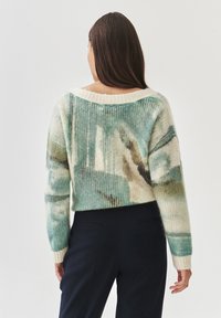 Frau mit langen braunen Haaren, die einen lockeren, mehrfarbigen Strickpullover und dunkle Hosen trägt, mit dem Rücken zu einem schlichten Hintergrund gewandt.