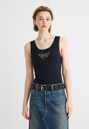 Jeune femme portant un débardeur noir avec un graphisme de papillon et une bordure en dentelle, une jupe en jean bleue et une ceinture noire à œillets, debout devant un fond blanc.