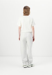 Persoon met schouderlang bruin haar in een wit T-shirt, grijze joggingsbroek en witte sneakers, staand met de rug naar de camera op een effen witte achtergrond.