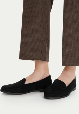 Czarne zamszowe mokasyny noszone z brązowymi, wzorzystymi spodniami o długości do kostek, na osobie stojącej na jasnym tle.