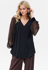 Czarny top z dekoltem w serek i luźnym krojem, z brązowymi, przejrzystymi, bufiastymi rękawami w groszki. Tkanina wydaje się gładka i lekka.