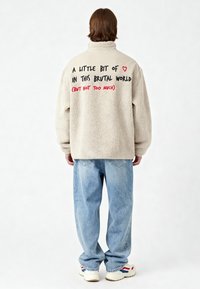 Veste en polaire beige à col montant, avec un texte noir et rouge au dos. Portée avec un jean bleu ample et des baskets blanches.