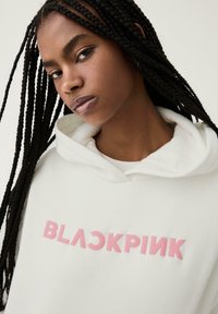 Haut à capuche blanc avec "BLACKPINK" en lettres majuscules rose pâle. Texture douce, capuche spacieuse et détails de couture visibles.