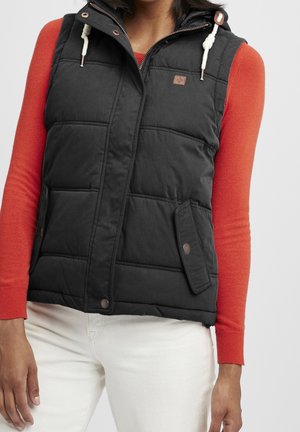 Kvinde iført en sort quiltet vest med forlommer over en orange langærmet trøje og hvide bukser, stående mod en ensfarvet baggrund.