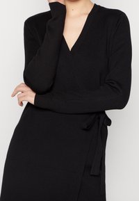 Svart omlottklänning med långa ärmar, V-ringning och knytning i midjan. Tillverkad av ett mjukt stickat material, med en elegant och figurnära design.