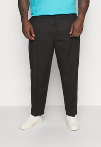 Man in zwarte rechttoe-rechtaan broek, witte sneakers en een blauw shirt, staand tegen een effen witte achtergrond.